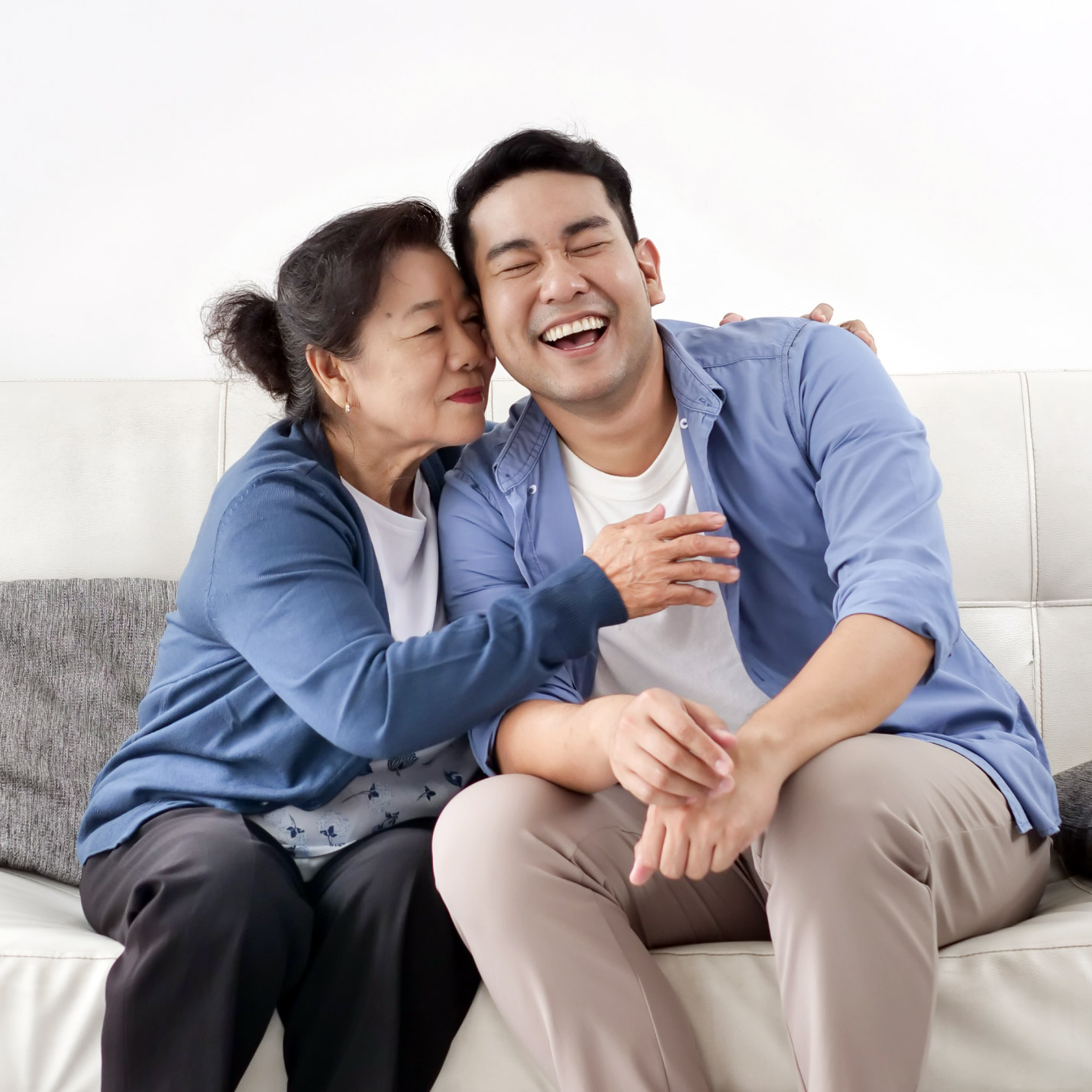 An elderly woman embracing her adult son who is laughing as they share a moment on her home couch together.